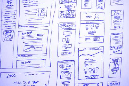Eine handgezeichnete Sammlung von Website-Layout-Skizzen, die auf einem Whiteboard angezeigt werden. Die Skizzen sind verschiedene Drahtgitterdesigns mit unterschiedlichen Anordnungen von Textblöcken, Bildern, Überschriften und anderen Webelementen. Die Skizzen sind mit blauer Tinte gezeichnet.